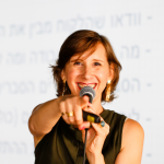 רונית כפיר מסתכלת לתוך מצלמה ומצביעה לכיוונך